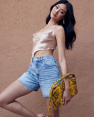 A model wearing a beige sleeveless handkerchief top, blue denim shorts, and holding a yellow snake print bag. Date Night. Shop Now.